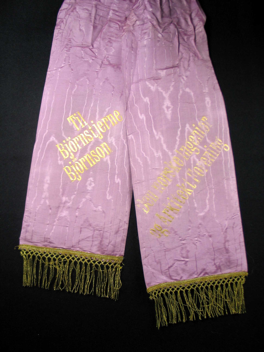 Begravelsessløyfe i lilla mønstret silke med frynser og tekst i gull.
