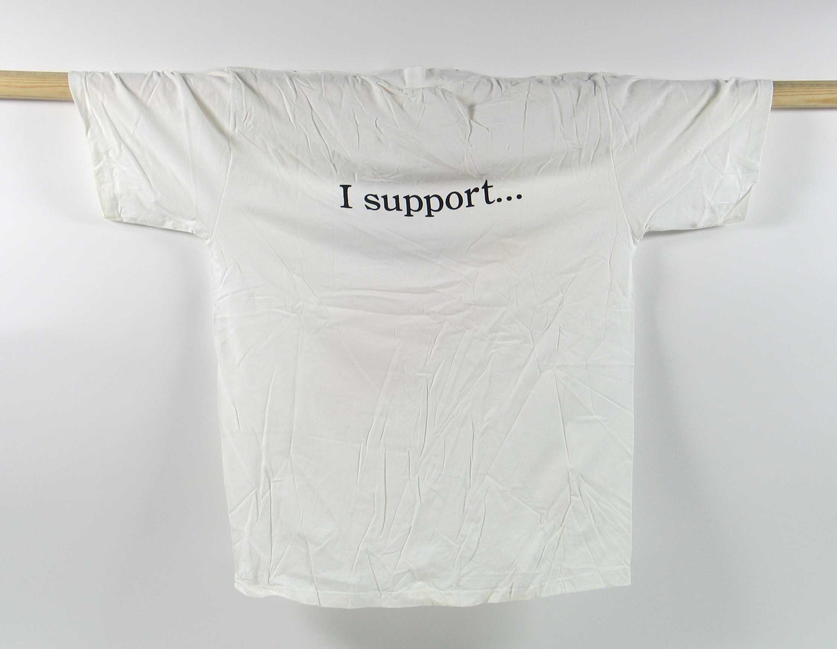 T-skjorte med hvit som hovedfarge. Størrelse x-large. På fremsiden er det en logo for OLYMPIC AID. I Logoen inngår et piktogram av Fakkelmannen som leier et barn i armen. På ryggen av skjorten står det skrevet: I support...