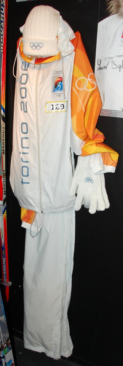 Hvit og oransje overtrekksdress bestående av jakke og bukse. Motiver av de olympiske ringene på ermer og på ryggen av jakken.