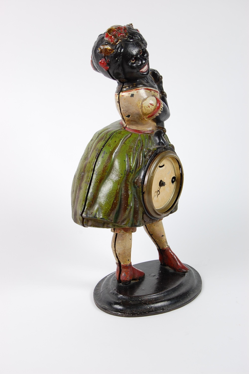 Støpt og malt figur formet som en afrikansk jente som bærer en klokke. Figuren har oval sokkel. Klokken er satt inn foran på skjørtet.