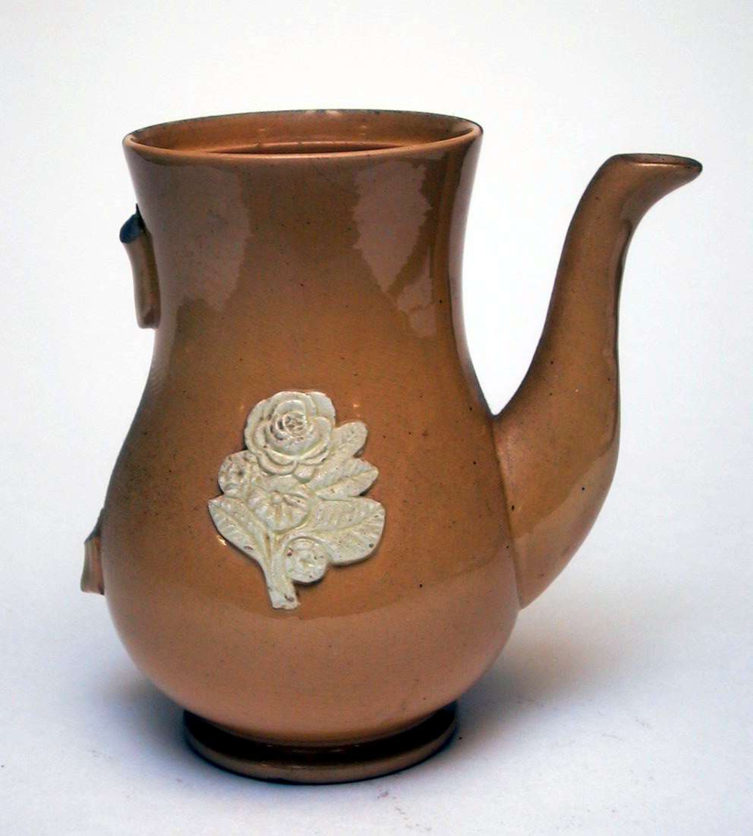 Kaffekanne av gul keramikk med påsatt blomsterdekor. Dekoren er elfenbenshvit. Lokket og hanken mangler.
Kannen har ingen produksjonsmerker. 