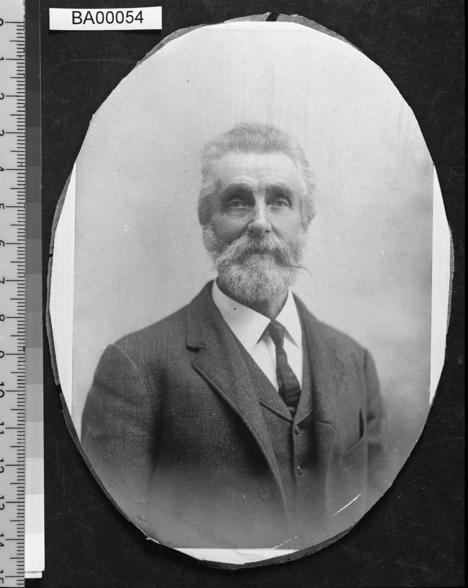 Portrettfotografi av eldre mann med bakoverbørstet hår og skegg med snurrbart;  kledd i mørk dress med vest og hvitt skjorte med slips. Han ser direkte på betrakteren.