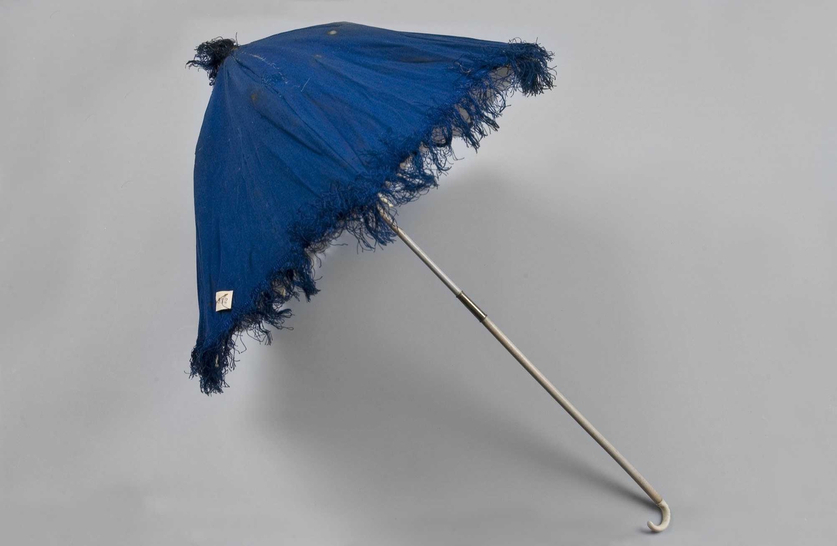 Liten blå parasoll med frynser langs kanten og på toppen. Foret med hvit silke. Spiler med pynteknotter. Stangen er todelt og låses med et messingbeslag, dekorert håndtak.