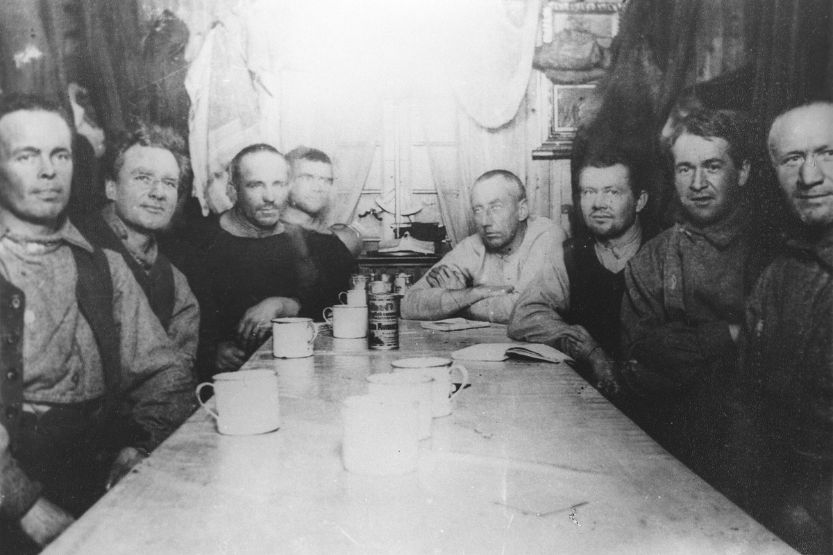 8 menn sittende rundt et bord. (Sydpolsekspedisjon). Roald Amundsen ved enden av bordet.