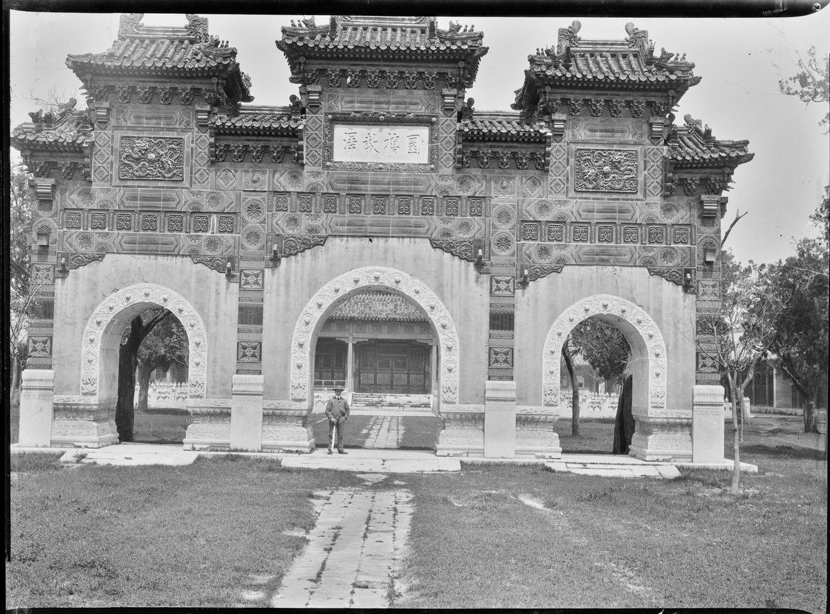 En turist under et steinmonument/portal med søyler og buede innganger. Trolig ved Sommerpalasset i Beijing.
