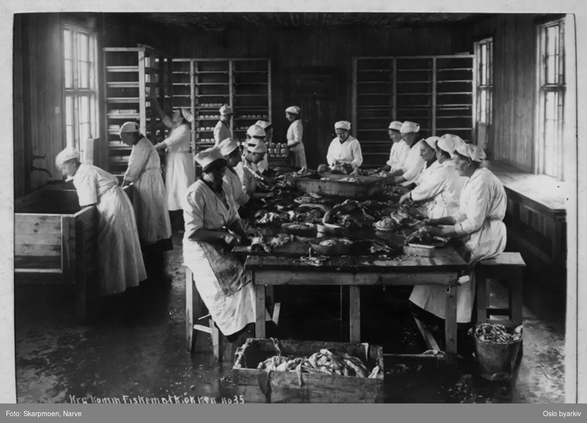 Mange kvinner i arbeid med å rense fisk, arbeider rundt et stort bord, kvinnene kledd i hvite frakker og skaut, store skyllekar, høye hyller med brett med fiskemat.