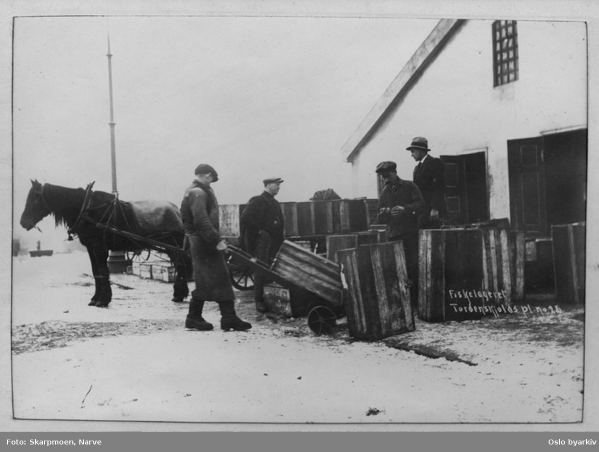Hest og vogn, menn med håndkjerrer, kasser. Lagerbygning på Tordenskjolds plass.