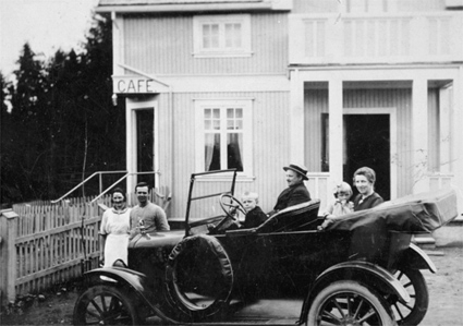 FORD T 1923-25 MODELL, BIL MED KALESJEN NEDE, HARALD HANSSEN MED FAMILIE I BILEN, FORAN HANSENS BUTIKK OG KAFE VED BRENNERIET I LØTEN. T-FORD. 
Harald Hanssen sitter bak rattet i bilen. Ved siden av sitter sønnen Jarle Hanssen. Bak sitter kona Olea Hanssen, med dattera Hjørdis Olga Hanssen, gift Hornseth på fanget. Foran bilen står Lovise Rasmussen, gift Tønsetengen sammen med Petter Søberg. 