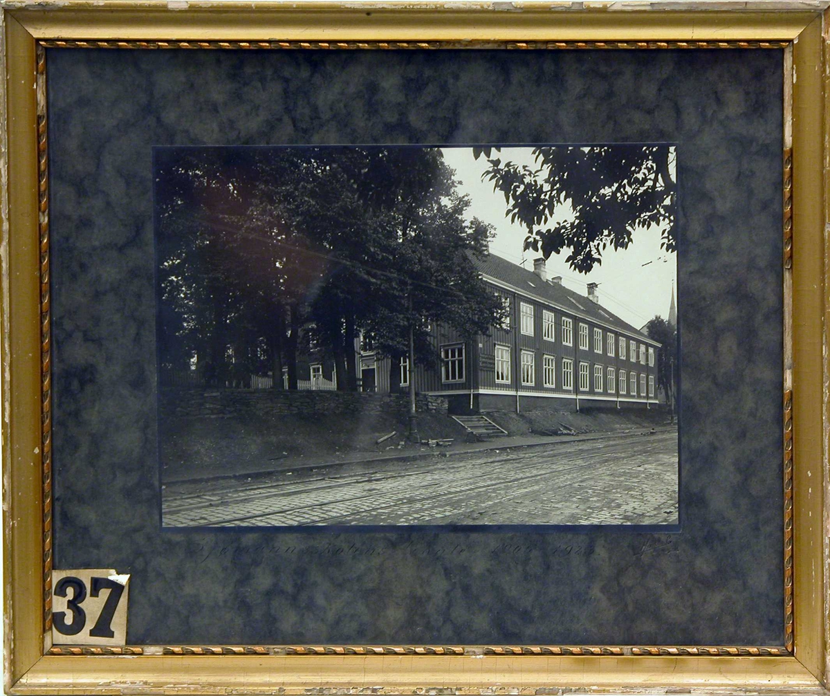 Fotografi av en større trebygning ved brolagt gate med trikkeskinner.  
Trondhjems Offentlige Sjømandsskole 1905-1923 ved Kongensgt. 93.