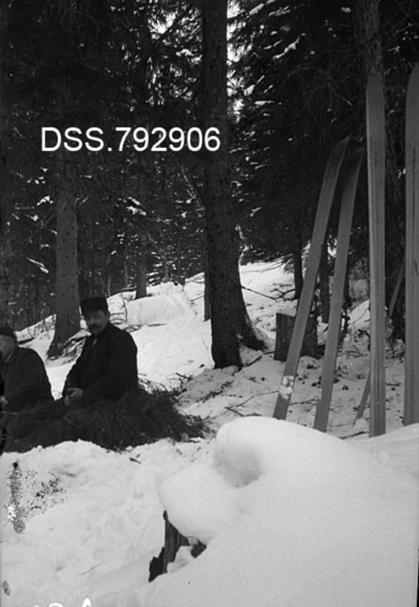 Rast under befaring i granskog, skogforvalter Hans Godtfred Støre og dr. Thrap-Meyer sitter på benk av granbar, sistnevnte er delvis vekklipt.  Oppreiste ski til høyre i bildet, granskog i bakgrunnen. 