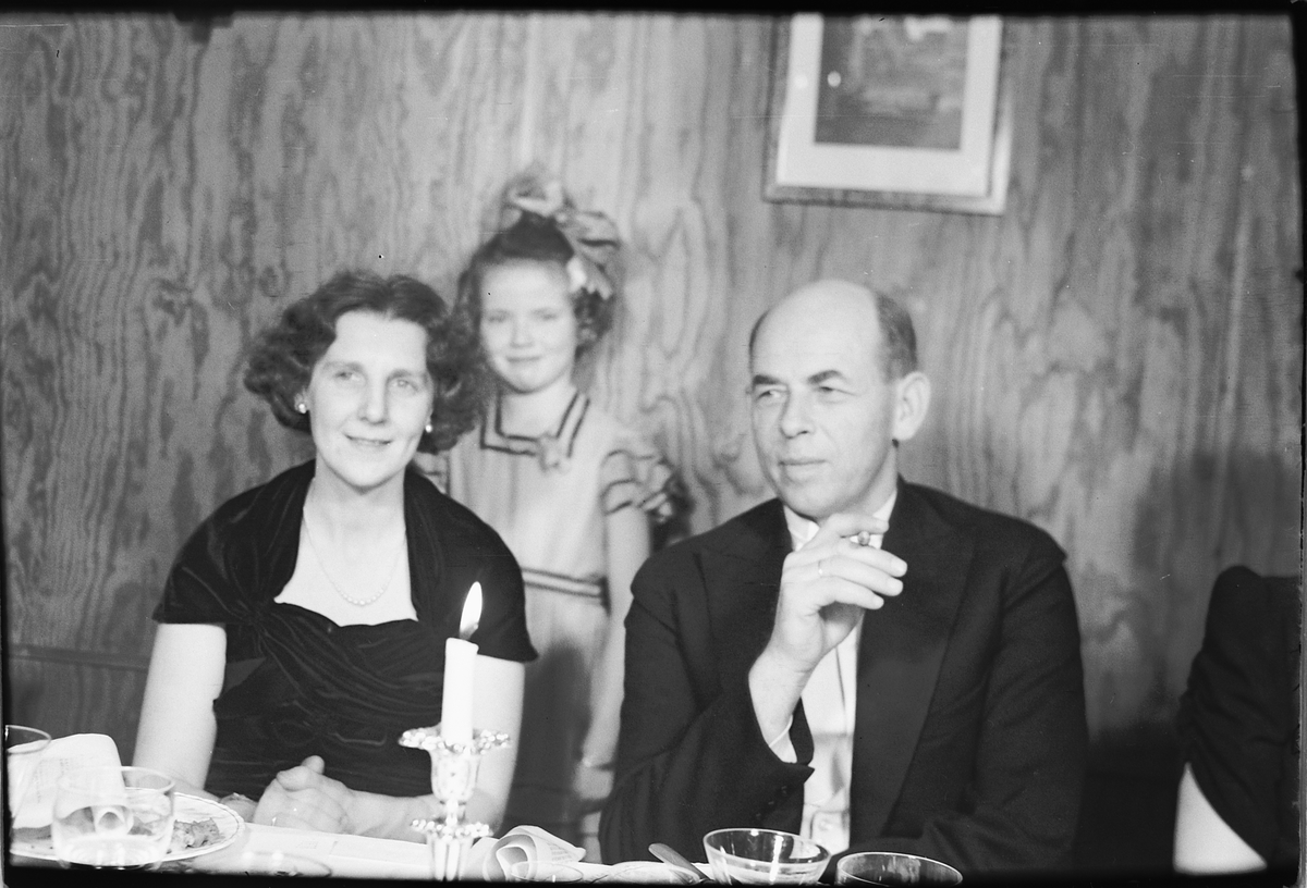 Arendal, sølvbryllup. Ukjent kvinne og mann sitter til bords. Jente med sløyfe står bak dem. 