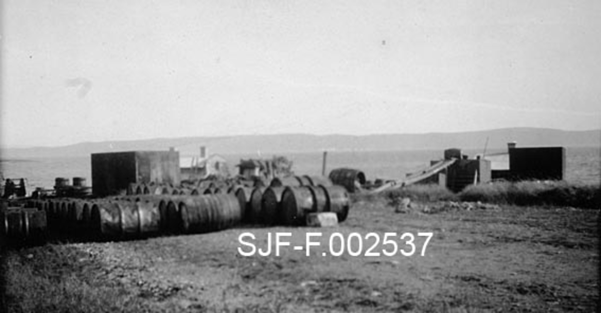 Fra Børsholmen i Asker, der Schwencke & Co's Eftf. i 1924 bygde et lite industrianlegg for framstilling av bek og andre tjærebaserte produkter. Fotografiet er tatt på ei flate på den øvre delen av holmen, som tydeligvis ved denne anledningen ble brukt som lagerplass.  Det som ble lagret her var framfor alt fat (tønner).  Sammen med tønnene lå det imidlertid også en diger, rektangulær stålbeholder (jfr. SJF-F. 002530).  Bakenfor og nedenfor den nevnte lagerflata skimter vi flere av de små bygningene som inngikk i dette industrianlegget.  Mot horisonten ser vi åsene på den andre sida av fjorden. 