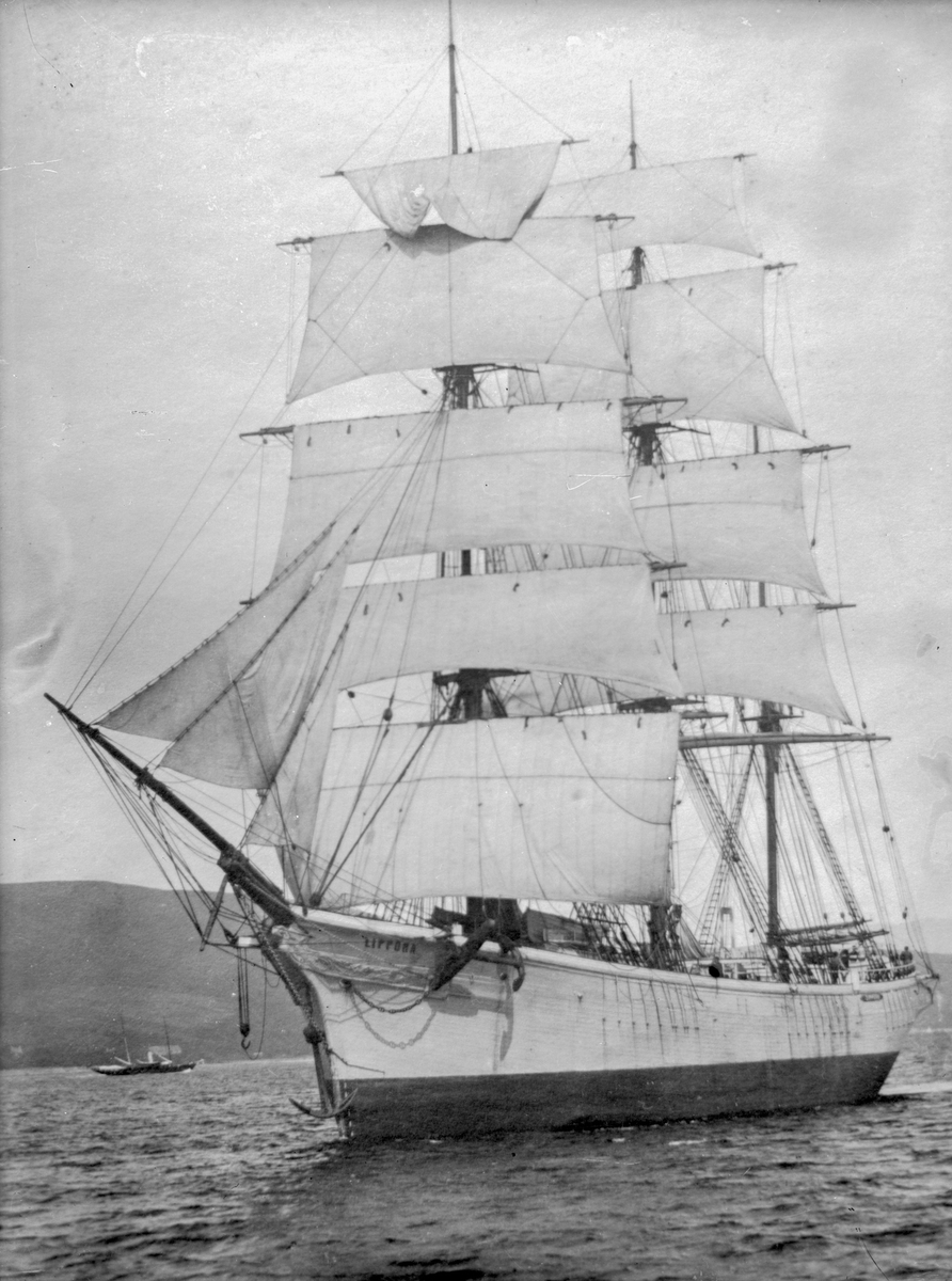 Fullriggeren "Zippora" for fulle seil langs kysten, med liten båt og høye fjell i bakgrunnen.