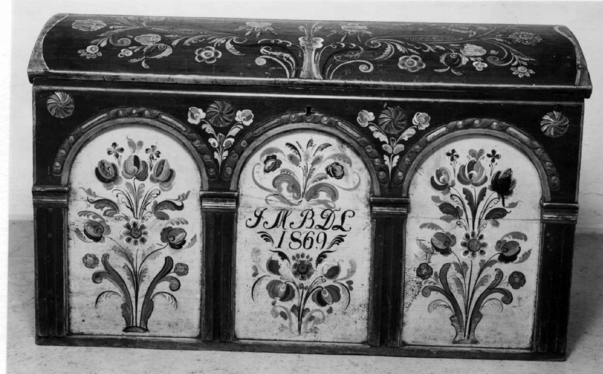 Kiste med buet lokk og rosemalt. På forsiden tre arkader, kortside ådret.