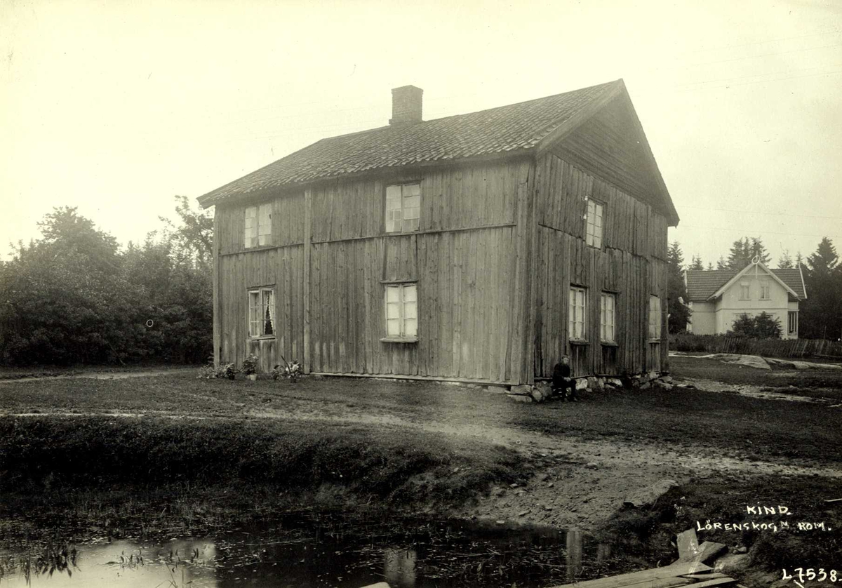 Kind, Lørenskog, Nedre Romerike, Akershus. Gammelt hus, mann sitter mot veggen, nyere hus i bakgrunnen.