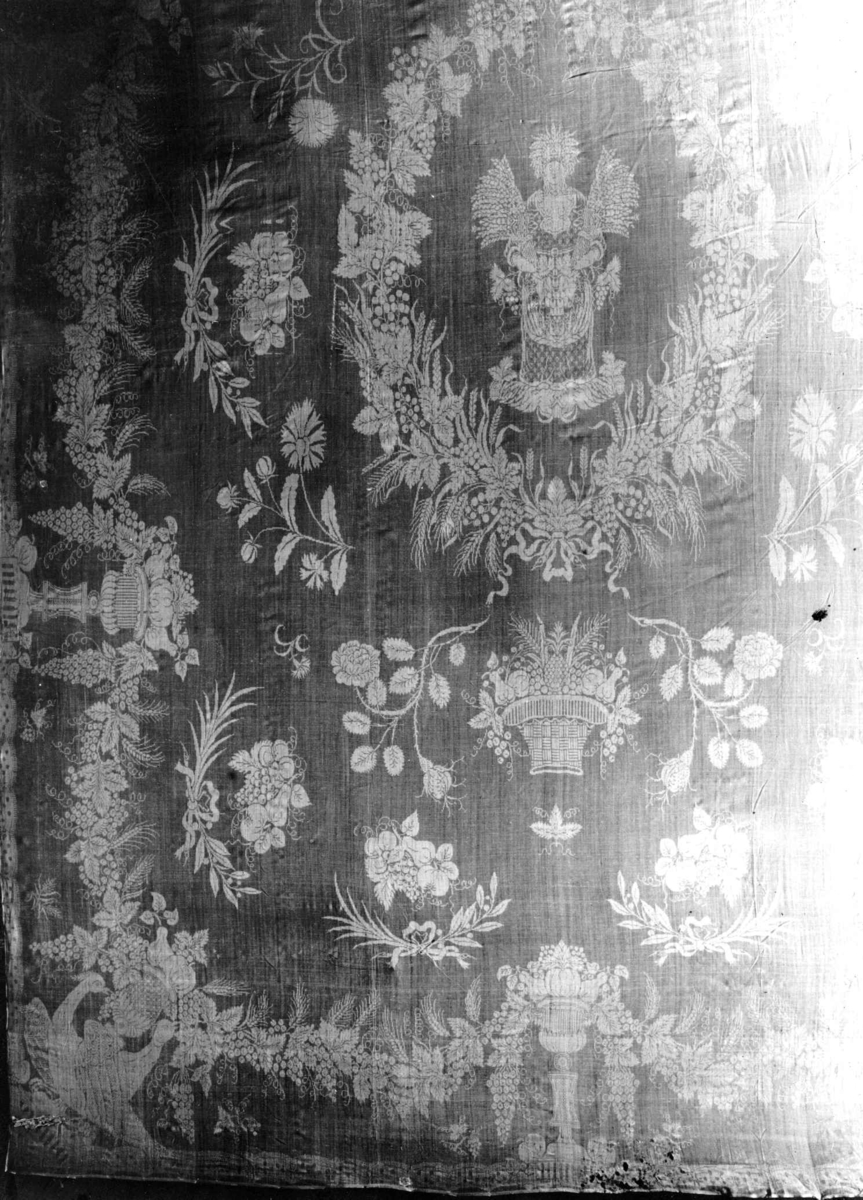 Detalj av hvit damaskduk med mønster av kone med kornnek omgitt av bladkrans og blomsterbuketter. Tyskland 1700-tallet.
