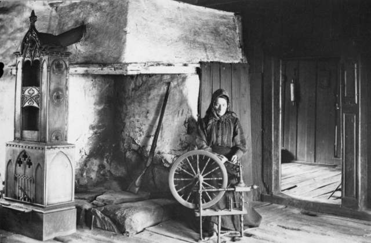 Kvinne sitter ved rokk foran grua i stua. Gevær inne i peisen, etasjeovn på venstre side, med ovnsrør inn på pipa.
