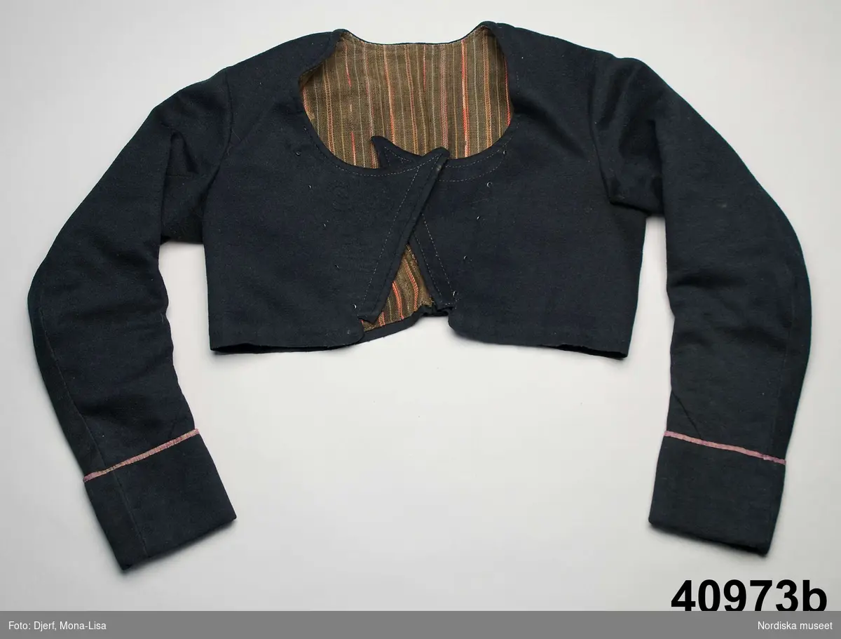 Huvudliggaren: Dräkten "begagnades till början av 1840-talet, linnet av senare årtal."
a.1-2 bindmössa m. stycke
b. tröja längd 35 cm, ärml. 46 cm
c. överdel
d. förkläde
e. kjol
f. 1-2, skor
g. 1-2, bindmössa m. stycke


Kvinnotröja, kort modell av svart kläde,  kallad midjetröja, 2 fram/sidstycken, 2 bakstycken som avsmalnar i ett litet skörtveck mitt bak, nederkanten råklippt och ofållad. framstyckena går omlott med snibb, det högra framstycket har en laskad cirkeldekor mitt fram. Dubbelknäppning med hakar och hyskor fästade genom tyget, isydd lång ärm formad med 2 sömmar, 9,5 cm brett ärmuppslag kantat med lila sidenband. Helfodrad med smalrandig halvbomullskypert med smala ränder på brun botten, de röda och vita ränderna i ullgarn, varpen gråbrunt lingarn, troligen en återbrukad kjol. Tröjan handsydd.
Se Greta Hedlund: Dräkt och kvinnlig slöjd i Ovansjö socken sid 116-119, 1981 där liknande tröjor beskrivs.
/Berit Eldvik 2010-11-09