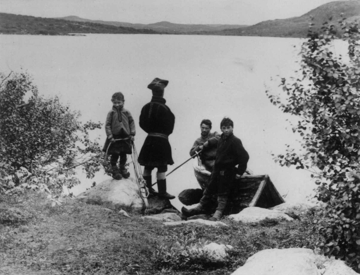 Fire samer ved en robåt i strandkanten, Sør-Varanger, Finnmark, ca. 1900. Gutt med fiskegarn?
