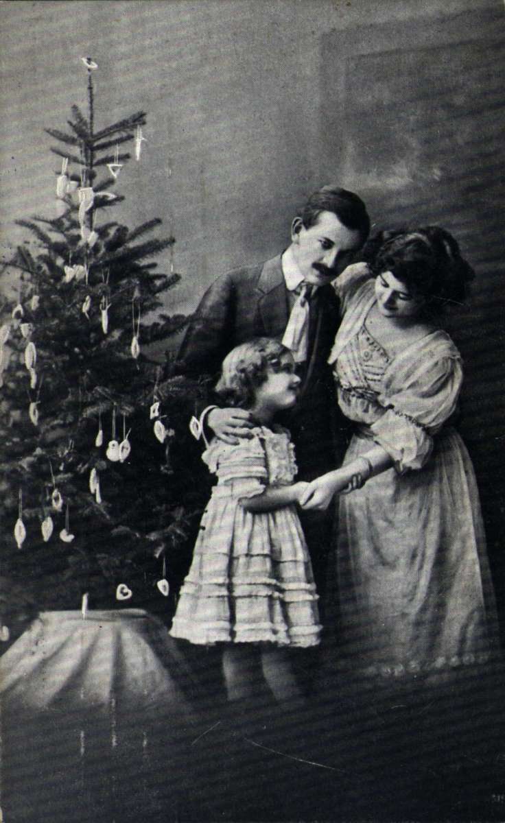 Julekort. Jule- og nyttårshilsen. Fotografi, svart/hvitt. Mor, far og datter ved siden av juletreet. Stemplet 23.12.1905.