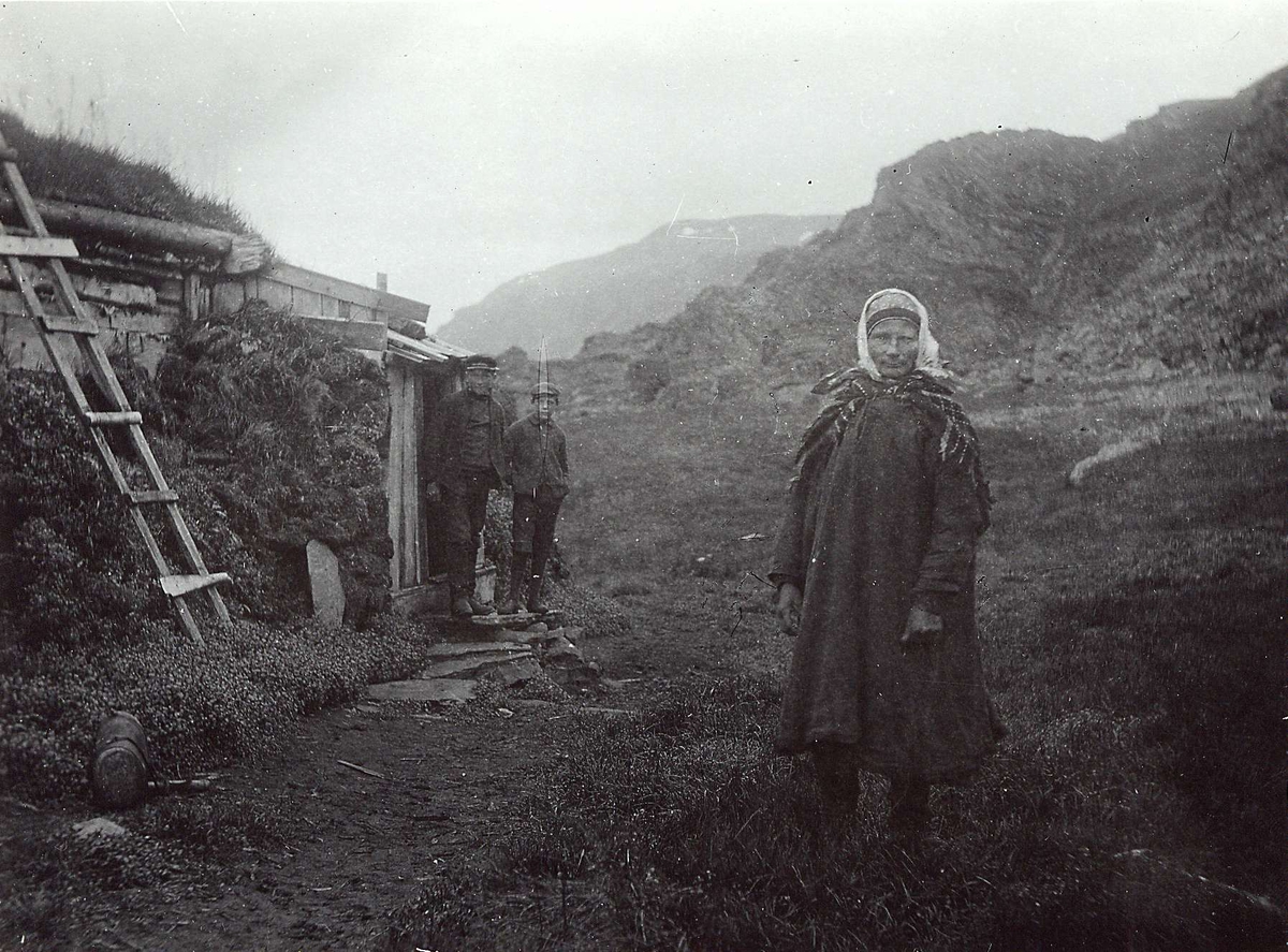 Kvinne i samisk drakt, Ellen, stående foran inngangen til gamme, Båtsfjord. Gutt og mann står i bakgrunnen, rett foran døra inn. Stige står lent mot gammen. 
Del av serie fra en forskningsreise i Øst-Finnmark 1909.