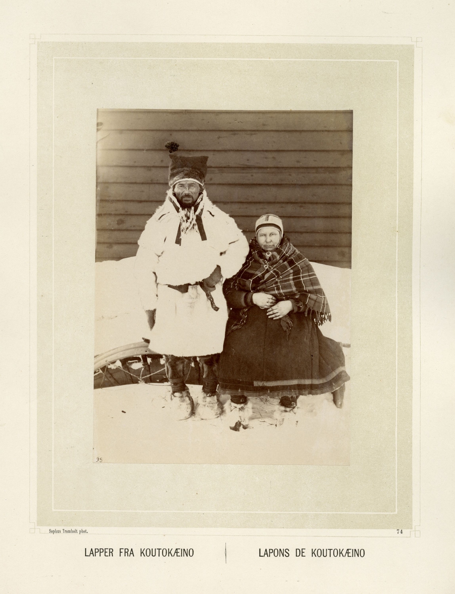 Samisk par fra Kautokeino sitter på baksiden av en pulk. De er kledd i pesk, kofte, sjal og skaller.
