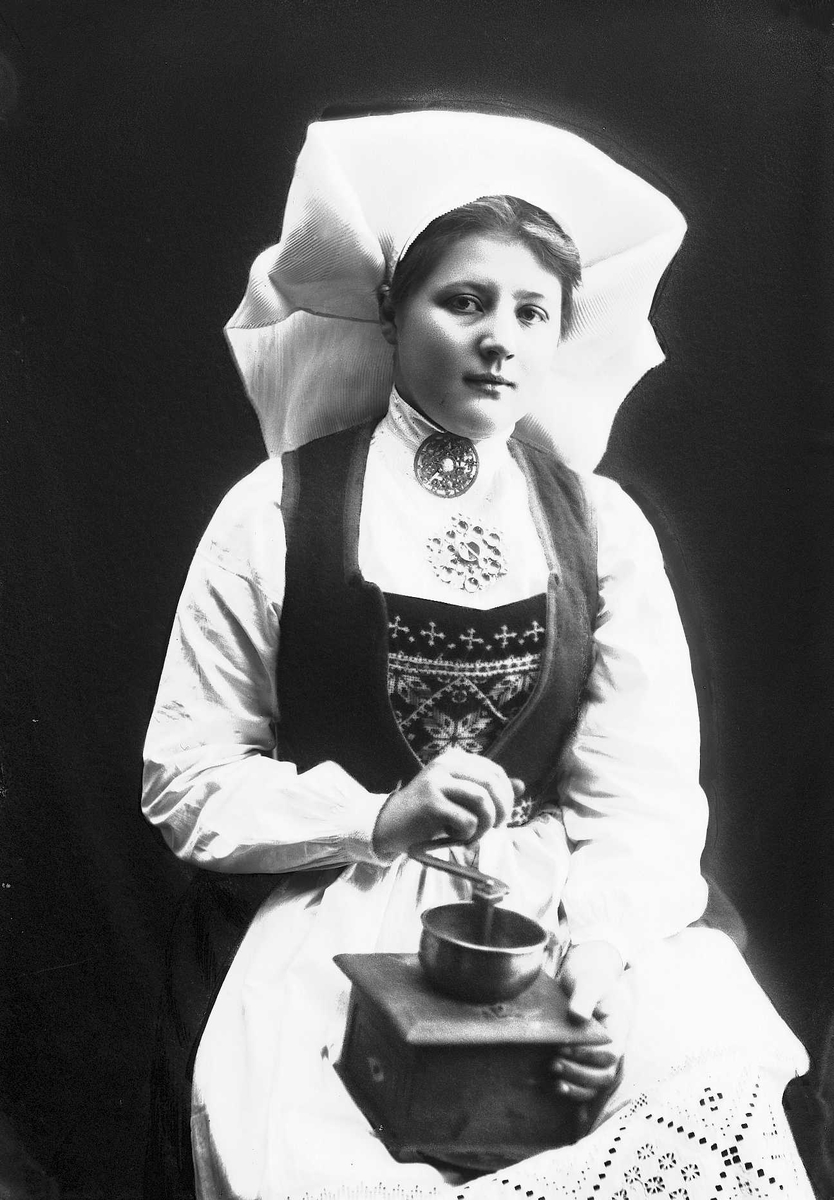 1/2 portrett av kvinne i drakt og skaut, tatt i fotografens studio. Hun sitter og holder en kaffekvern på fanget. Nøytral bakgrunn.