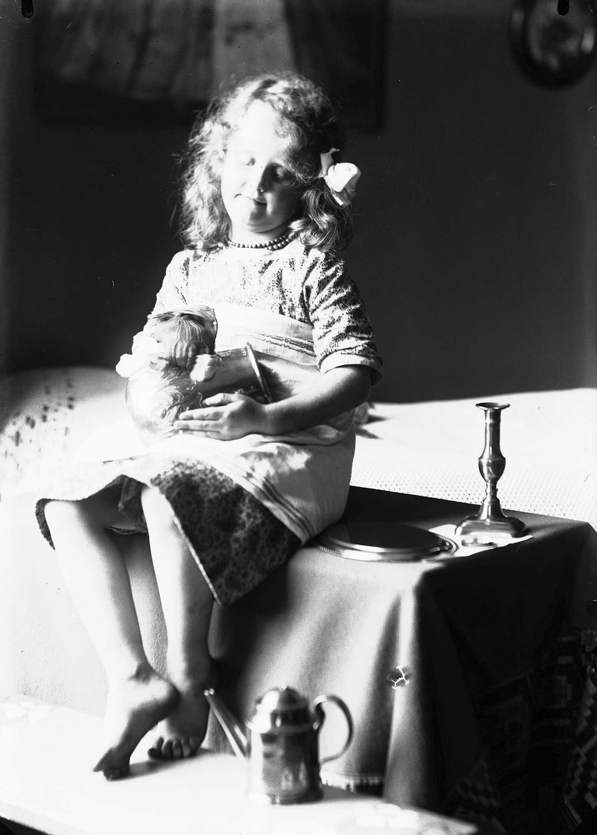 Pike sitter på bord med metallvase på fanget som hun pusser. På bordet ligger tallerken, lysestake og på benken foran står en kanne. Piken har perlehalskjede og blomst bak øret. Seng i bakgrunnen.
