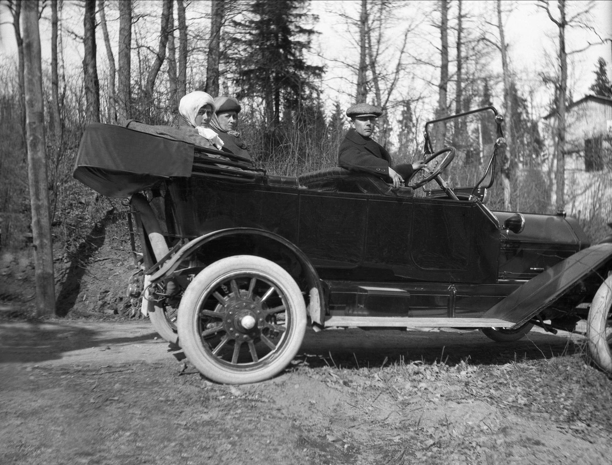 En mann sitter bak rattet på en parkert åpen bil med to damer i baksetet. Robsahm og Lund.