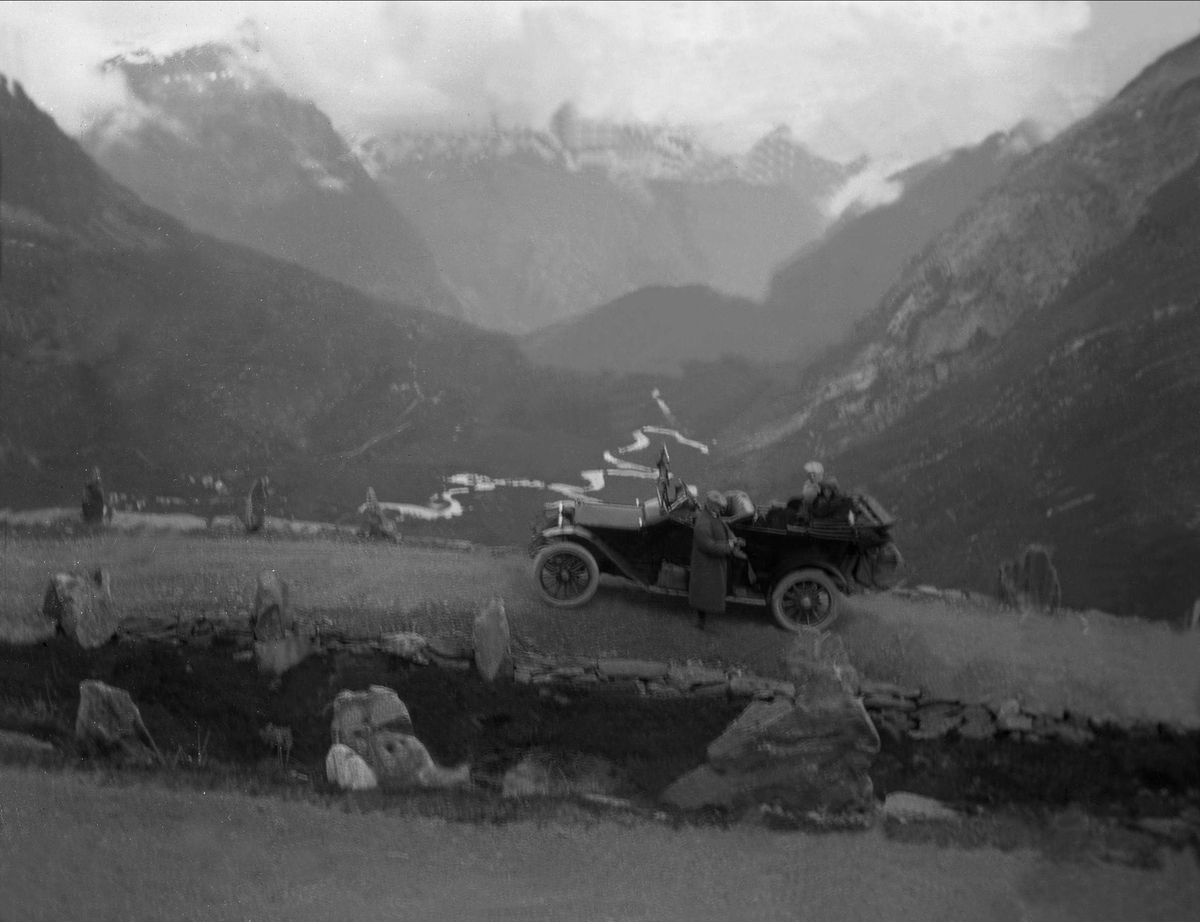 En bil med to personer i baksetet står parkert på en grusvei med utstikt i bakgrunnen. En mann står ved siden av bilen. Robsahm og Lund.