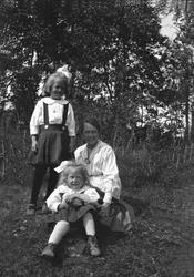 En kvinne og to barn poserer på gresset i kjoler. Robsahm og
