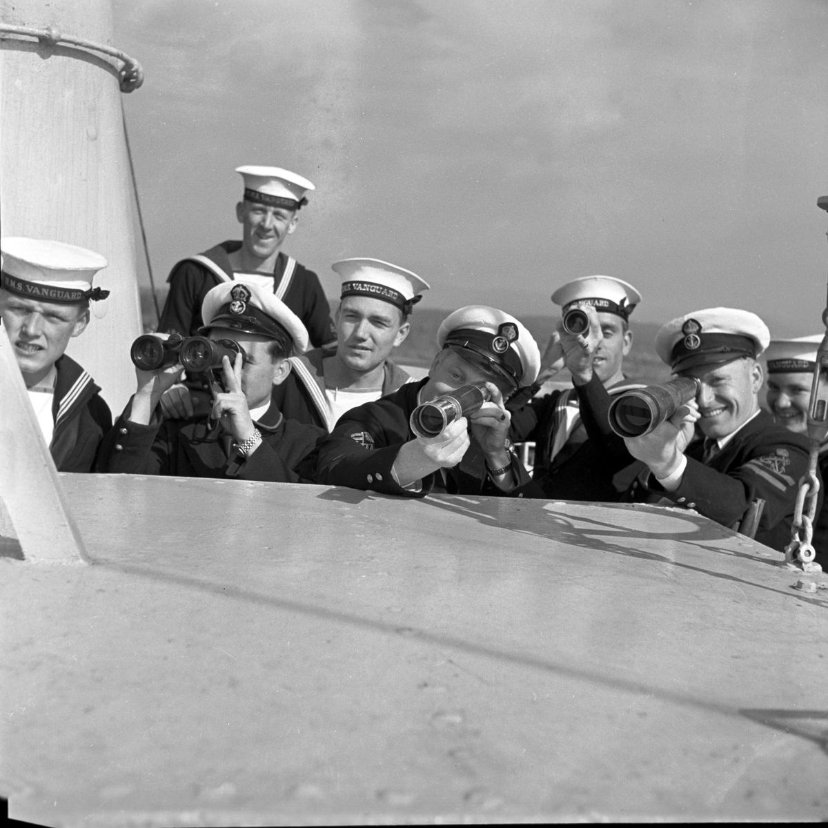 Serie. Det engelske slagskipet HMS Vanguard på flåtebesøk i Oslo. Parade med musikkorps ombord. Noen av mannskapet står med kikkerter og en pusser kanoner. Fotografert 21. juni 1954.