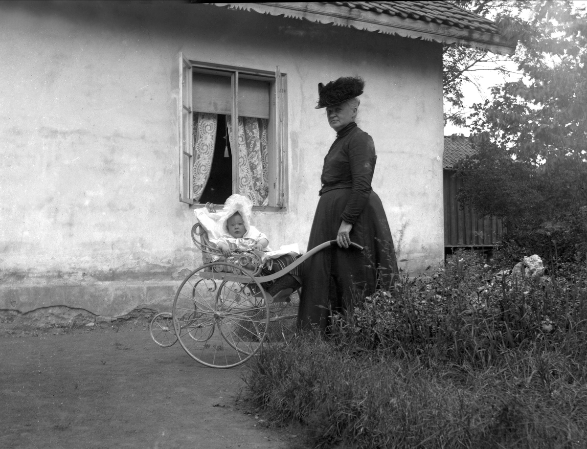 Eldre kvinne med barn i barnevogn, i hage utenfor pusset murhus. Avfotografering(?)