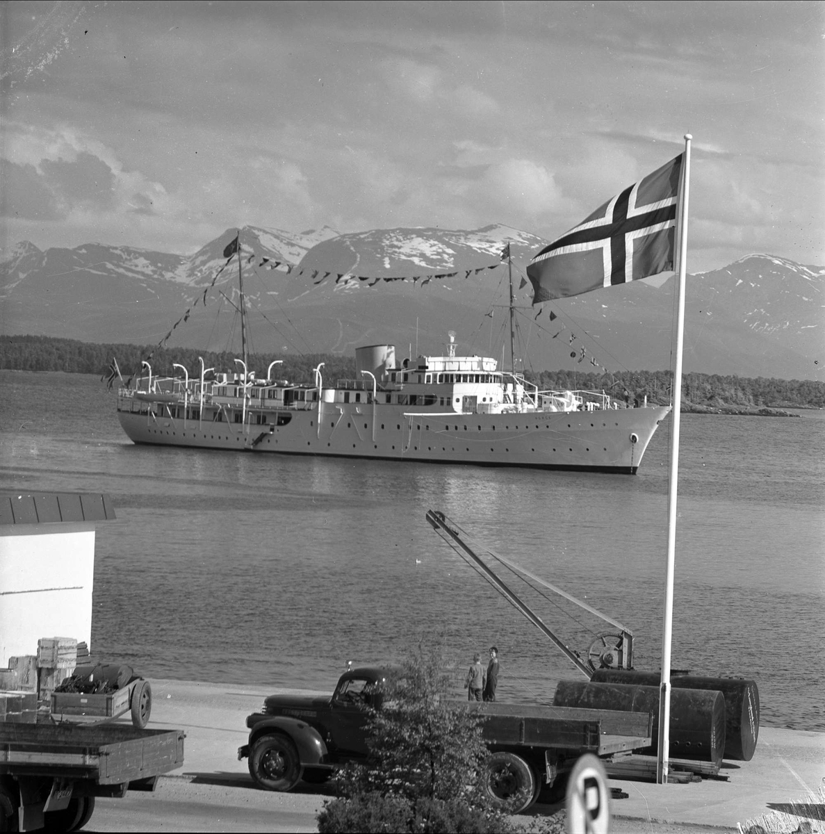 Kongeskipet Norge kommer inn fjorden med fjell i bakgrunnen. Antakelig fra Ålesund på signingsferden til kong Olav. Lastebiler på kaia med flagg,