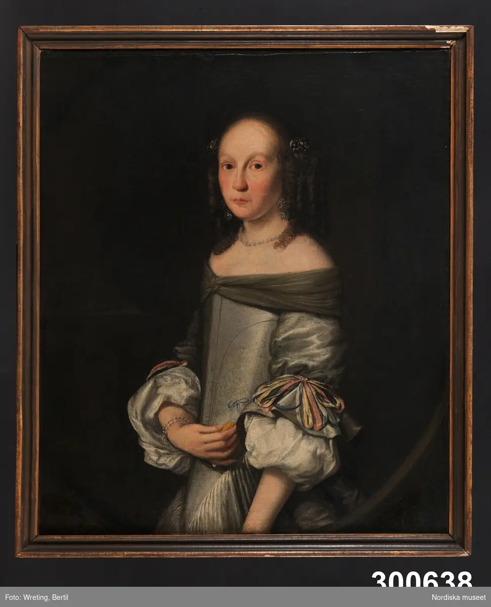 Porträtt av ung kvinna, midjebild fas vänster. Mörkbrunt hår med korkskruvslockar, gråvit sidenklänning med åtsittande liv.