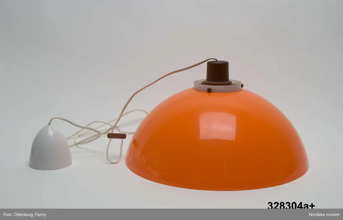 a+ = lampskärm
b = glödlampa 
a+) Taklampa av plast, för elektrisk belysning. Halvklotformad skärm, kupa, av orange plast. Lamphållare av brunt trä håller skärm och vit plastcylinder som inuti skärmen omger glödlampan lodrätt från lamphållaren ned till lampkupans nederkant. Sladd- och takfäste av vit plast, sladden avslutas med kontakt. Klistermärke på takfästets kopp med texten: "MADE IN SWEDEN BY LUXUS VITTSJÖ DES UNO o ÖSTEN KRISTIANSSON". Ingjutet i koppens insida: "LUXUS MADE IN SWEDEN".
b) Glödlampa, vitt halvgenomskinligt glas, päronformad, skruvfäste av vit metall. Text på toppen "PHILIPS CLASSICTONE 60W 230V". Tillverkningsår sannolikt 2004. Längd 10 cm.

Givaren köpte taklampan vid flytt från Malmö till Stora Mossen, Bromma 1959 och har använt den som taklampa över köksbordet fram till 2004.
/Maria Maxén 2006-01-23