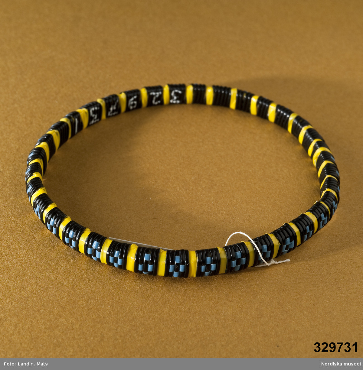 Tvärrandigt runt armband i gult och rutor i blått mot svart botten. Vävt av tunna plastband. Afrikanska influenser.
/Zingoalla Rosenqvist 2009-02-05