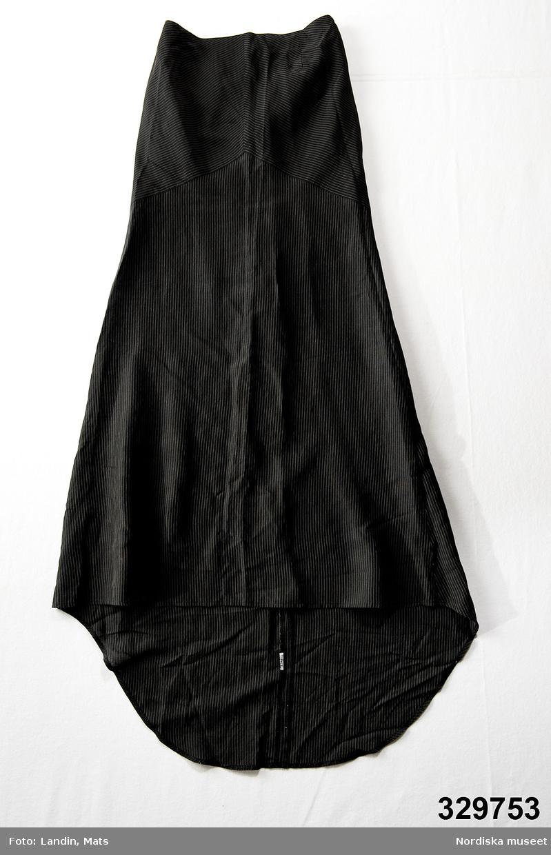 Fotsid kjol i tunt svart tyg med kritstrecksränder, lätt utställd med släpande  bakstycke. Höftpartiet är skuret på snedden av tyget. Insyningar upptill i fram- och bakstycket, dragkedja mitt bak. Infodrad upptill med grönt tyg.
/Zingoalla Rosenqvist 2009-02-05