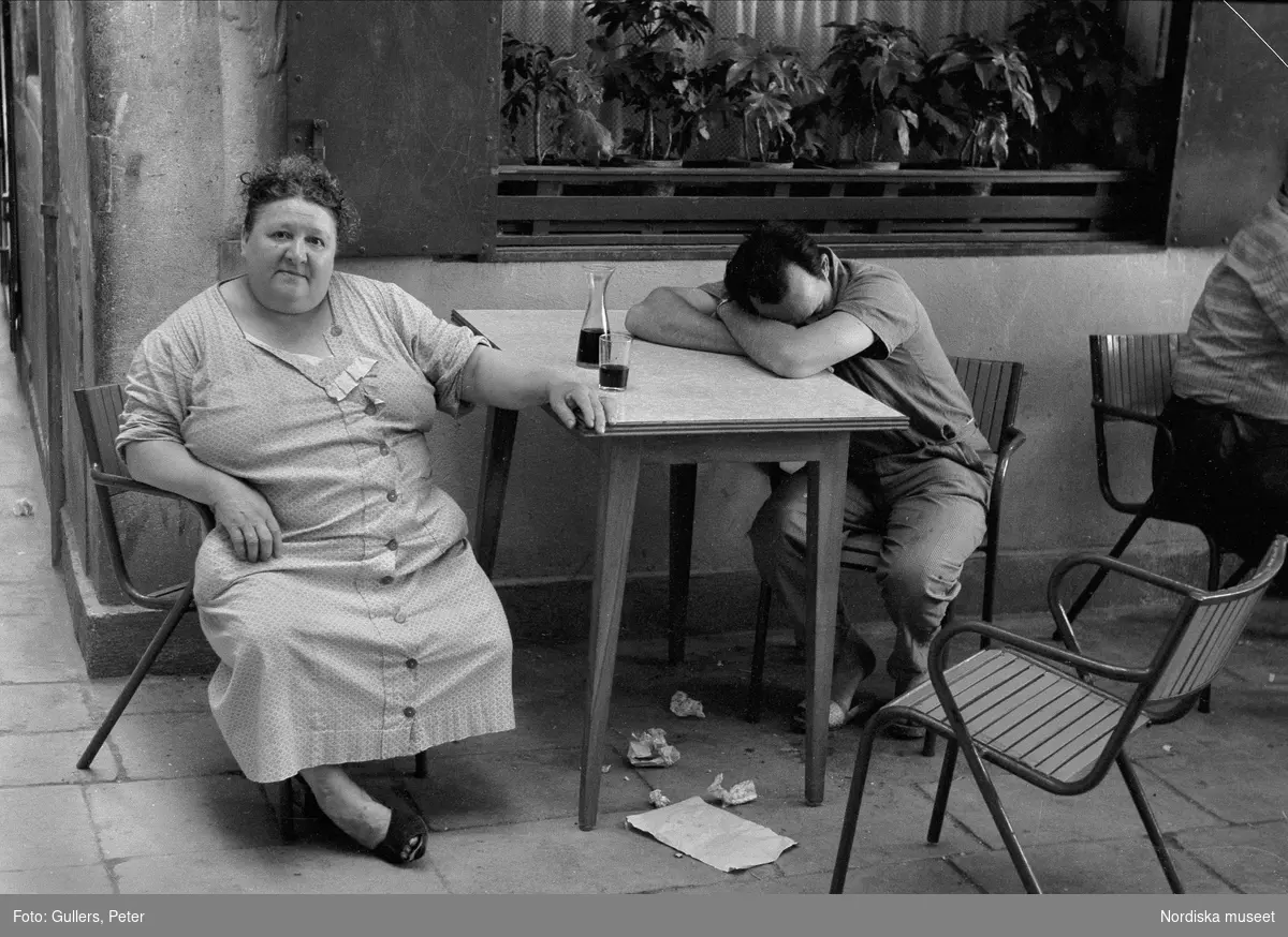 En kvinna och en man på en uteservering. Framför kvinnan står en karaff och ett glas med vin. Mannen lutar sitt huvud mot armarna som han korsat på bordet. Italien.