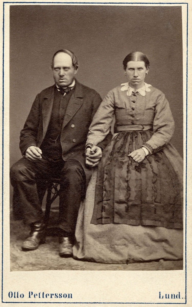 Ett medelålders par poserar sittande på stolar, de håller varandra i handen. från Löddeköpinge, Harjagers härad i Skåne. Inv.nr. 18518P.