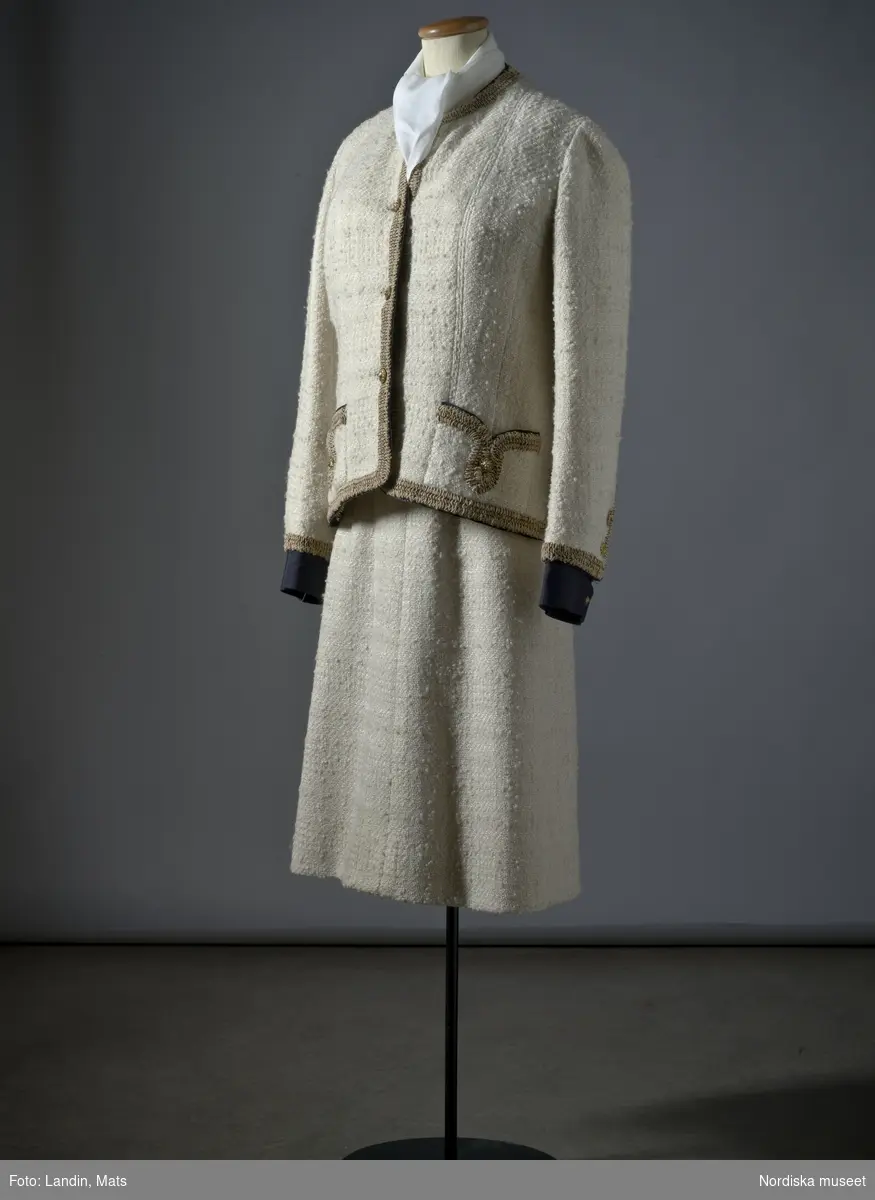 Dräkt av vit ylletweed med bandgarnering av svart ripsband och beige-bruna snörmakeriband. Inköpt hos modehuset Chanel i Paris 1961. Nordiska museet inv nr 283341.