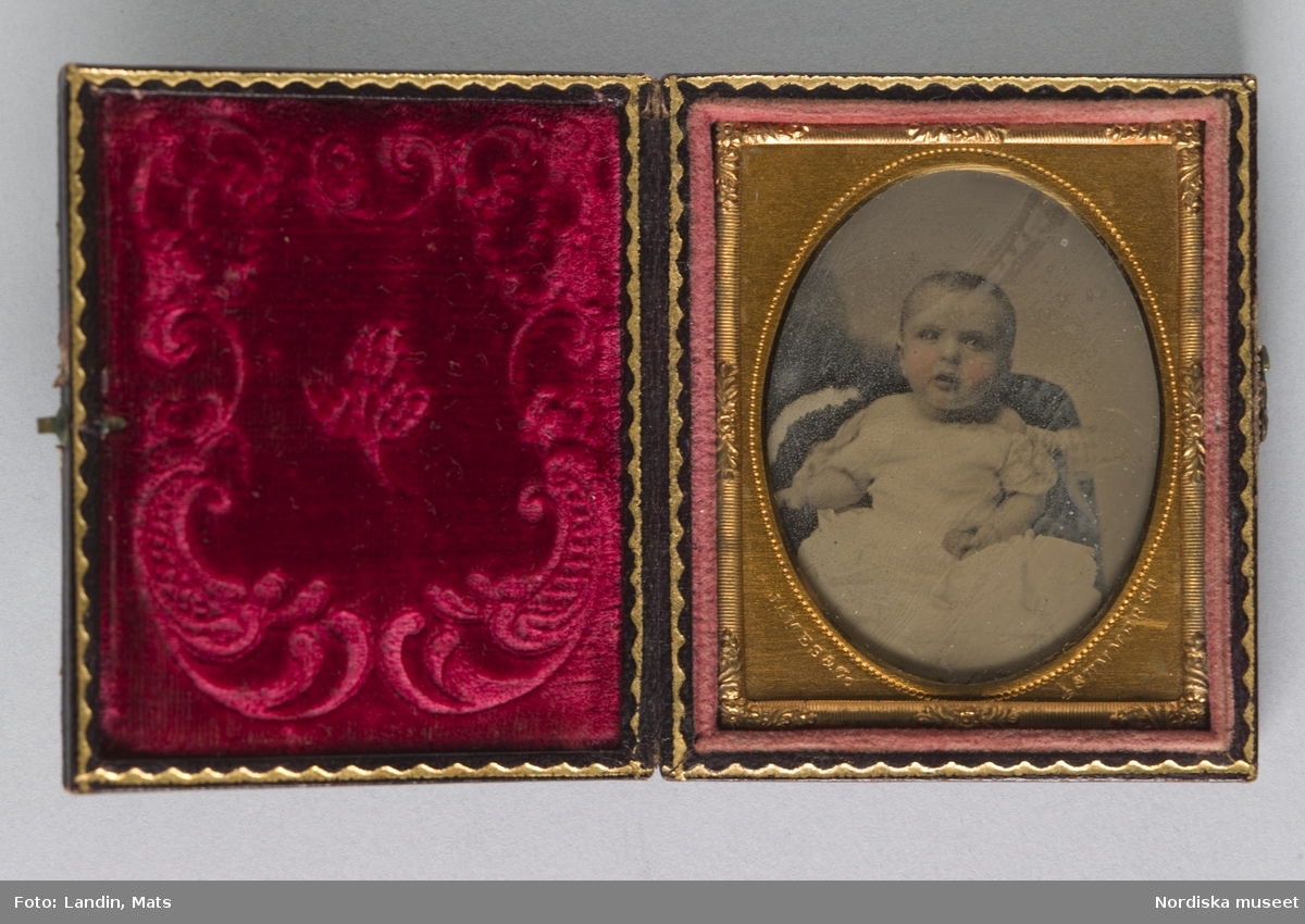 Porträtt av ett litet barn i vit klänning.Kolorerad Ambrotypi i förgylld metallram inpassad i fodral med sammetsfoder. Instansat i passepartouten: "James & Co, Summer St, 1860."
Nordiska museet inv.nr 279402
