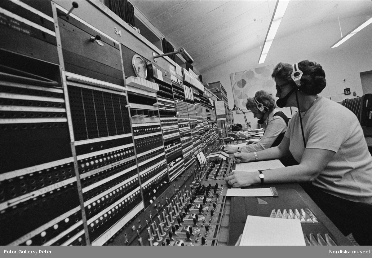 Telefonväxel. Några kvinnor arbetar med hörlurar och mikrofon framför en panel med knappar och reglage.