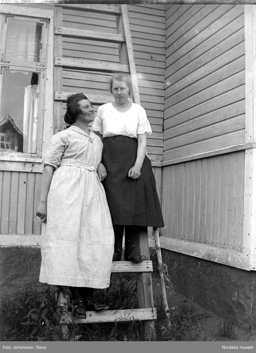 Två kvinnor på en stege som står lutad mot en husvägg.
