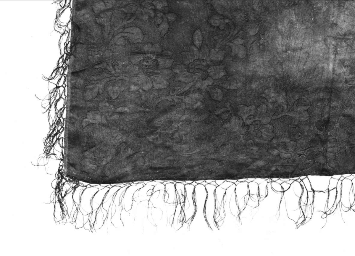 Huvudkläde av svart mönstervävt siden. Blommönster över hela ytan. Svarta iknutna silkefransar. Fållad för hand i två sidor.