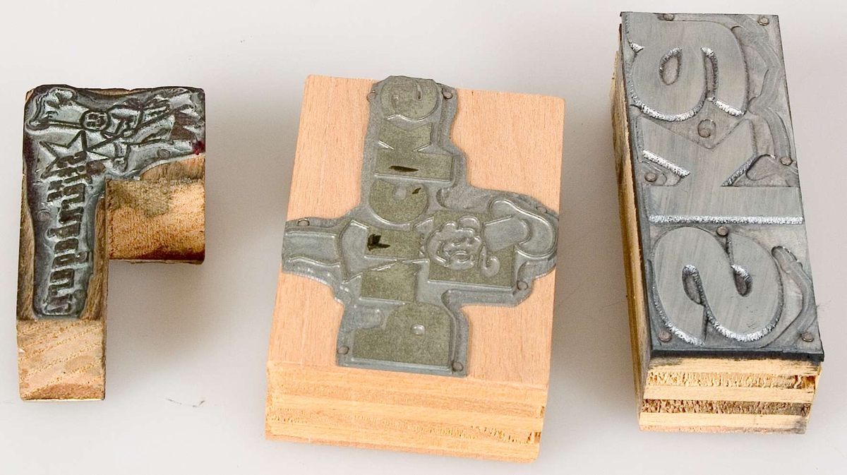 Tre klichéer av metall, olika storlekar och former, monterade på skiktlimmade klossar av trä.