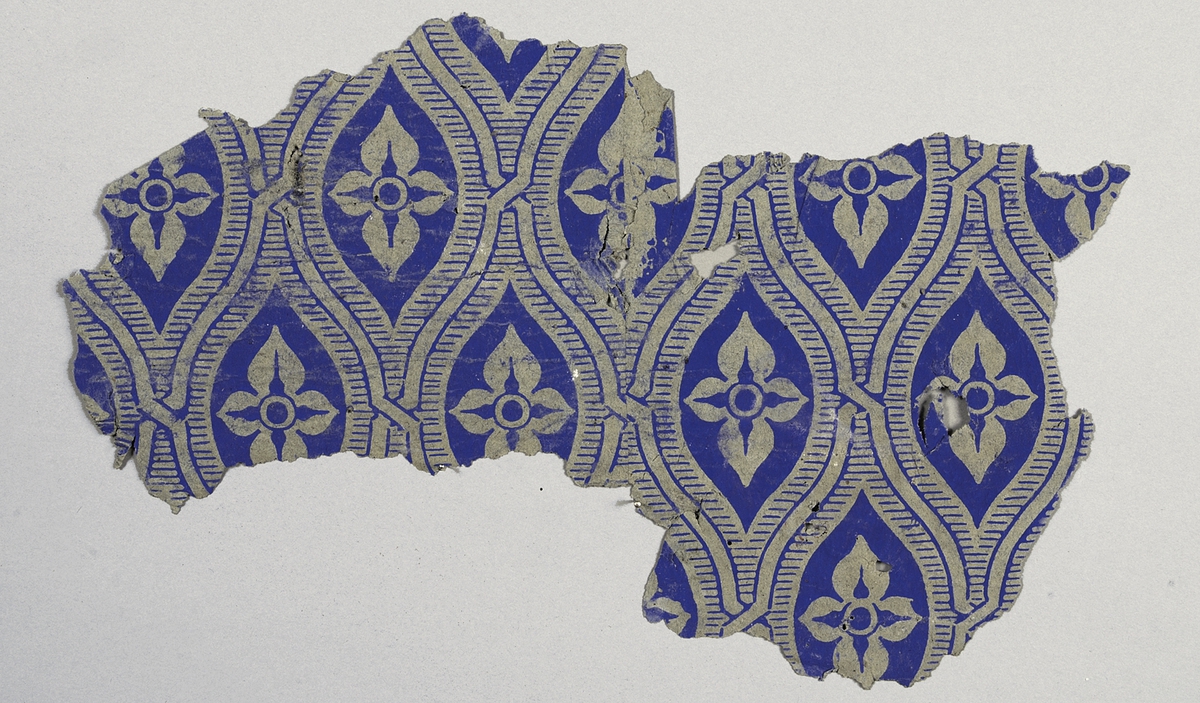 Tapetprovet (3 st. fragment) har brun grundfärg, med tryck i blått. 