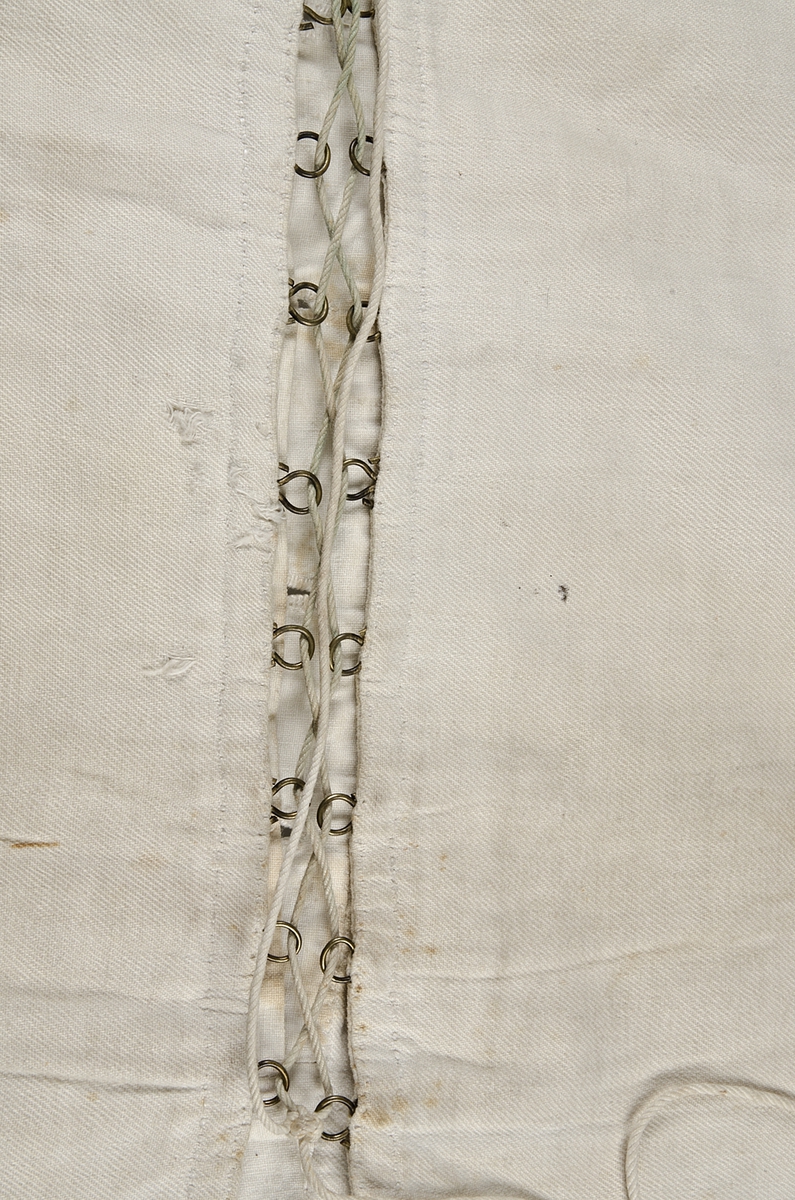 Snörliv, av vit bomull (kypert). Helt handsytt. Två delar som snörs samman bak. Snöret löper genom metallhyskor. Knäpps framtill med sex knappar (fem porslin och en metall). 1840-60-tal (nyrokoko).