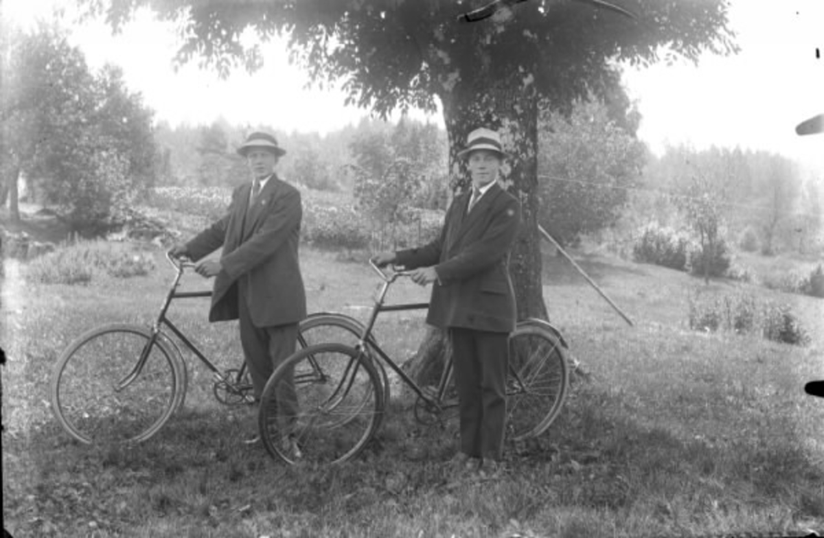 Gruppbild av två män med cyklar. Båda männen är klädda i hatt. De står vid ett träd i en hage. I bakgrunden skymtare en stenbro. Herman Nilsson och Erik Andersson (namnuppgifterna är osäkra).