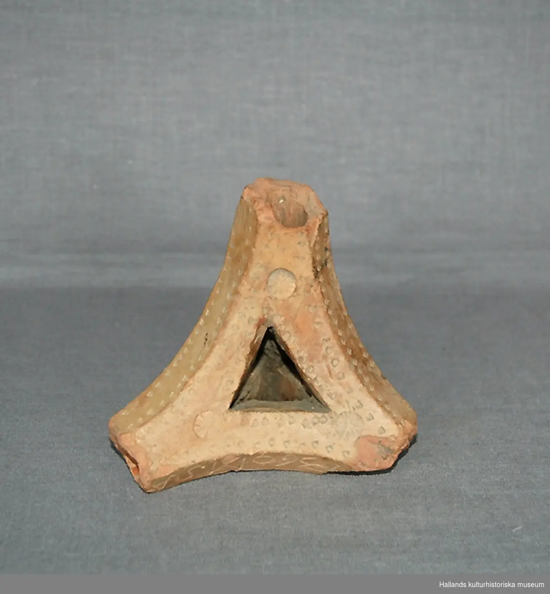 Ljusstake (fotangelstake) av bränt lergods. Trekantig form, består av fyra pipor. När en pipa används fungerar de övriga tre som fot. Dekorerad med ett tryckt mönster i lergodset. 
Märkt: "1360 MSA" (datering?)